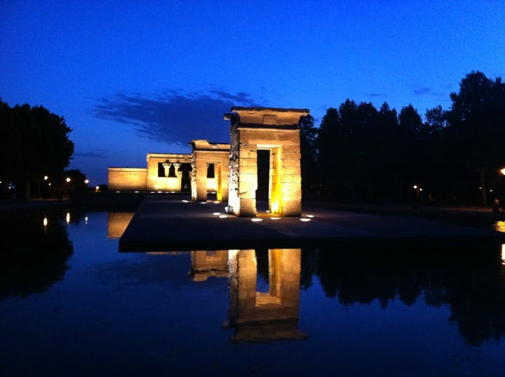 Upplysta gamla stenstrukturer återspeglas i en lugn vattenyta i skymningen, med träd och en djupblå himmel i bakgrunden.