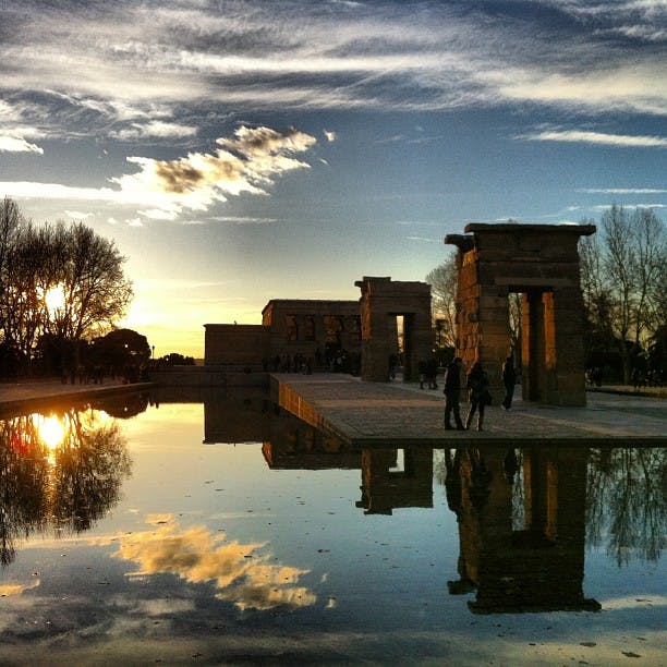 Antiche strutture in pietra che si riflettono in una piscina tranquilla al tramonto, con persone che passeggiano nelle vicinanze e un cielo parzialmente nuvoloso sopra.