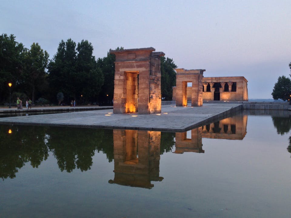 Forntida stentempel med upplysta pelare reflekterade i en lugn vattenyta, omgiven av träd i skymningen.