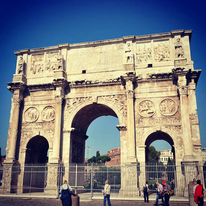 People walking near a large stone triumphal arch with intricate carvings and three arches, under a clear blue sky.