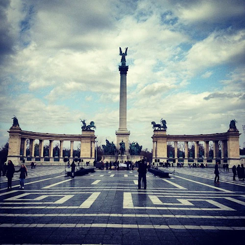 Ett stort torg med en hög pelare toppad av en staty, flankerad av två pelargångar med statyer; människor som går runt.