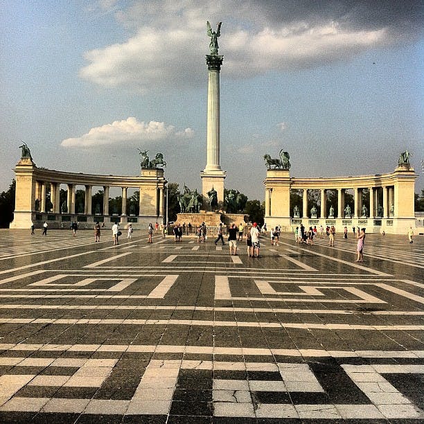 Folk går runt Hjältarnas torg med en hög pelare toppad med en staty, flankerad av två pelargångar med statyer.