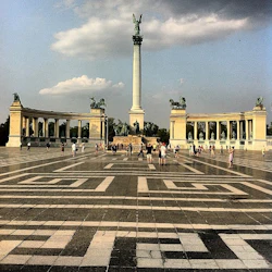 Folk går runt Hjältarnas torg med en hög pelare toppad med en staty, flankerad av två pelargångar med statyer.