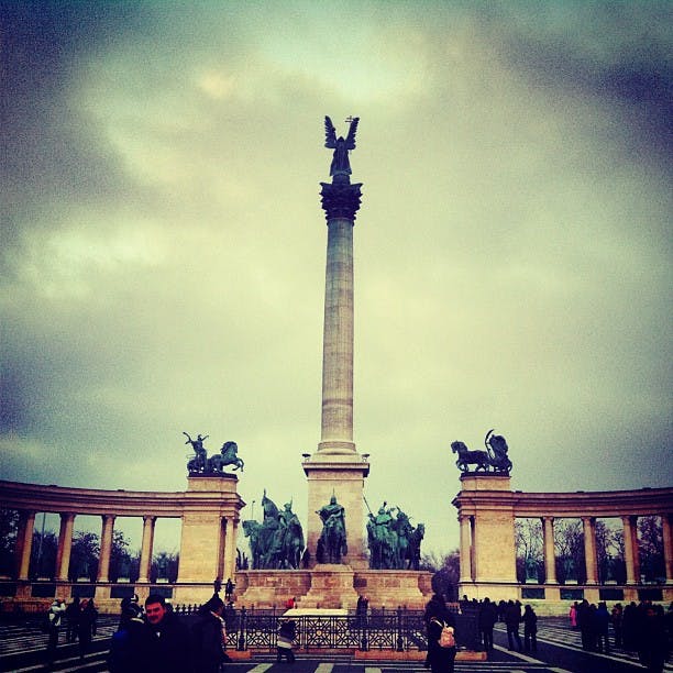 En stor staty med en ängel ovanpå en hög pelare, flankerad av statyer av hästar och ryttare, omgiven av halvcirkelformade pelargångar.