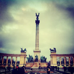 En stor staty med en ängel ovanpå en hög pelare, flankerad av statyer av hästar och ryttare, omgiven av halvcirkelformade pelargångar.