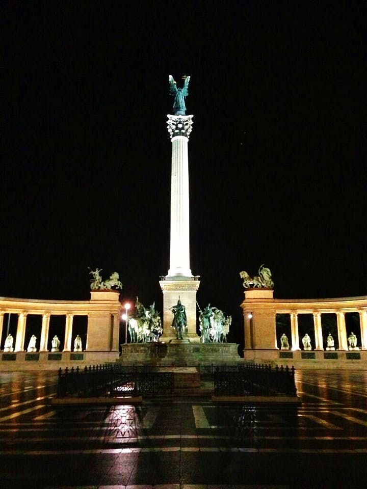 En upplyst pelare toppad med en staty står i mitten flankerad av statyer på en krökt arkad på natten.