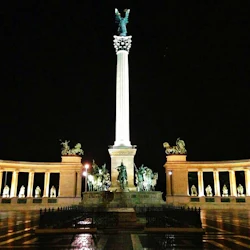En upplyst pelare toppad med en staty står i mitten flankerad av statyer på en krökt arkad på natten.
