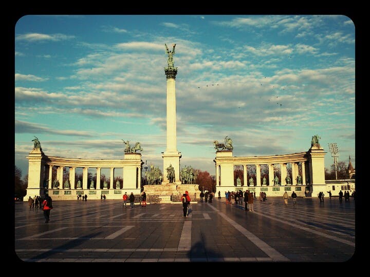Människor går runt ett stort torg med en hög pelare toppad av en staty, omgiven av halvcirkelformade pelargångar.
