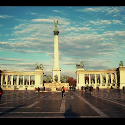 Människor går runt ett stort torg med en hög pelare toppad av en staty, omgiven av halvcirkelformade pelargångar.