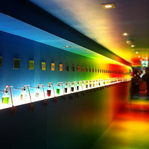 En lång hall med numrerade hyllor som visar glasbehållare fyllda med färgade vätskor, upplysta av levande, färgstark belysning.