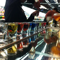 En rad färgglada shotglas på en bardisk med händer som förbereder drycker i bakgrunden.