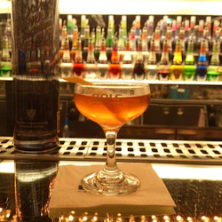 Ett cocktailglas fyllt med en bärnstensdryck, garnerad med en citron twist, sitter på en servett i en bar. Rader av färgglada flaskor finns i bakgrunden.