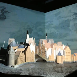 En detaljerad skalmodell av en historisk europeisk stad med olika byggnader, torn och en bro, mot en blå bakgrund.