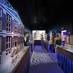 Människor i ett mörkt rum observerar detaljerade arkitektoniska modeller; golvet är täckt med sand och bakgrunden har en stadsbildsprojektion.