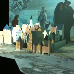 En detaljerad miniatyrmodell av en medeltida stad med olika byggnader ställs mot en stor bakgrund av spöklika, historiska figurer.