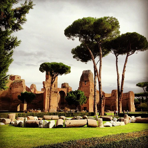 Höga träd omger antika ruiner och stenfragment på en grön gräsmatta under en molnig himmel.