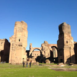 Tre personer står framför gamla, höga tegelruiner under en klarblå himmel.