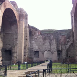 Massiva antika ruiner med stora valv, omgivna av stenmurar och järnräcken. Folk går runt och läser skyltar.