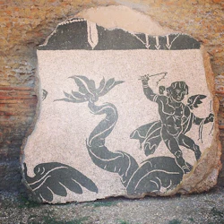 Forntida mosaik som visar en bevingad figur som håller i ett vapen, konfronterar en stor orm, ställd mot en grov tegelvägg.