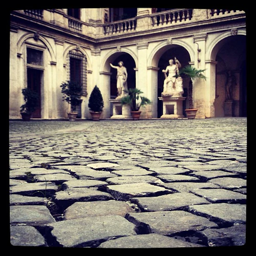 En kullerstensgård med krukväxter, statyer och välvda ingångar avbildas.