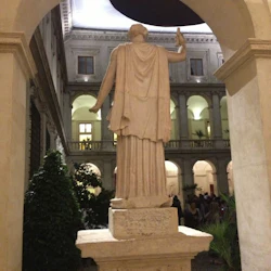 En marmorstaty, sedd bakifrån, står på en välvd innergård med krukväxter och människor i bakgrunden.