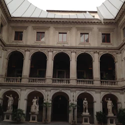 En historisk innergård med två våningar av valv, klassiska statyer och utsmyckade fönster under ett delvis täckt tak.