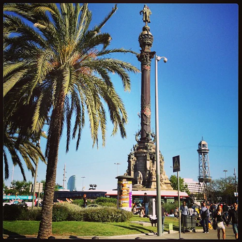 En hög palm står bredvid en stor, utsmyckad pelare med en staty ovanpå. Människor går runt på ett pulserande torg en solig dag.