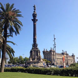 Högt monument med en staty på toppen, omgiven av grönska. Palmträd till vänster, historisk byggnad i bakgrunden. Klarblå himmel.