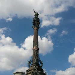 Högt monument utsmyckat med statyer, toppat av en figur som håller ett föremål, mot en blå himmel med spridda moln.
