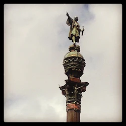 Staty av en man som pekar ovanpå en dekorerad pelare mot en molnig himmel. Kolumnen har utsmyckade mönster och figurer.