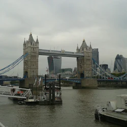 Tower Bridge över Themsen med moderna stadsbyggnader i bakgrunden, båtar på vattnet nedanför. Molnig himmel.
