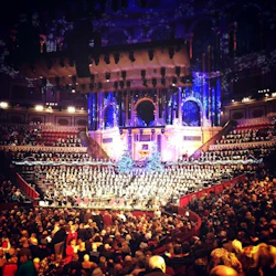 En fullsatt konsertsal med en stor kör på scenen, dekorerad med julgranar, som uppträder under färgglada ljus.