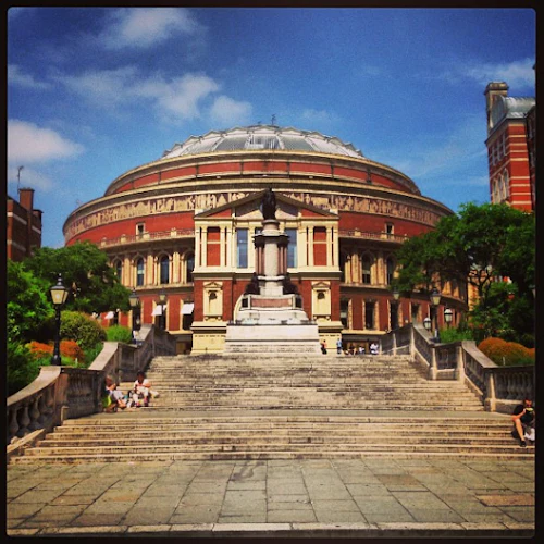 Människor sitter på trappor som leder till en stor, välvd byggnad med pelare och en staty i mitten. Träd flankerar trappan.