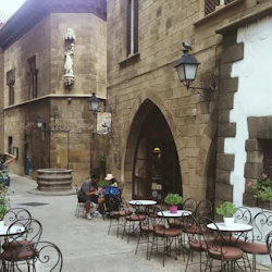 Utomhuscafé med järnstolar och små bord, beläget i en smal kullerstensgränd bredvid rustika stenbyggnader.